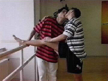 Same men kissing, one with leg on dance bar.