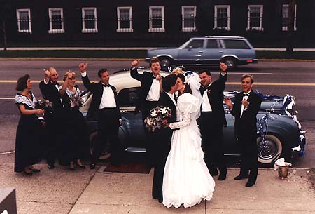 May 1991 - Wedding party cheers as Craig kisses Kimberly in front of blue classic car at their wedding