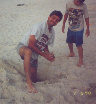 Jerry and James digging a hole on the beach.  Jerry's foot is buried in the hole and the ocean water is coming up through the bottom of it. 