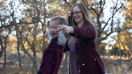 a mother stands behind a young toddler girl holding her hands for balance in a wooded nature area