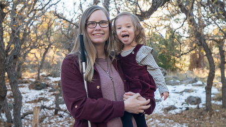 Brooke, a blind mother with glasses and a white cain, holds a blonde toddler girl in her arms as both smile at the camera. Medium shot from the waist up.