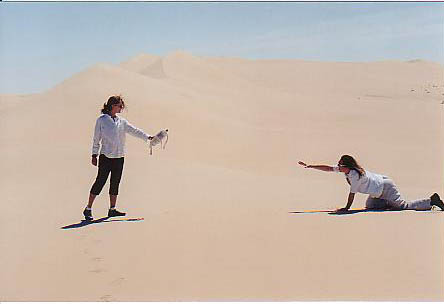 Donna crawls on hands and knees through the Scorching Sahara Desert towards Gina who is holding out a bottle of water