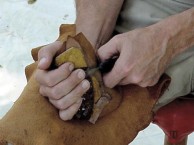 Picture of a Flint knapper chipping an arrowhead out of stone - he uses a leather pad on his leg and one in the palm of his hand for protection - he chips the stone in the palm of his hand
