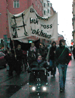 En funktionshindrad i permobil och en icke-funktionshindrad marcherar i ett demonstrationst�g framf�r en banderoll.