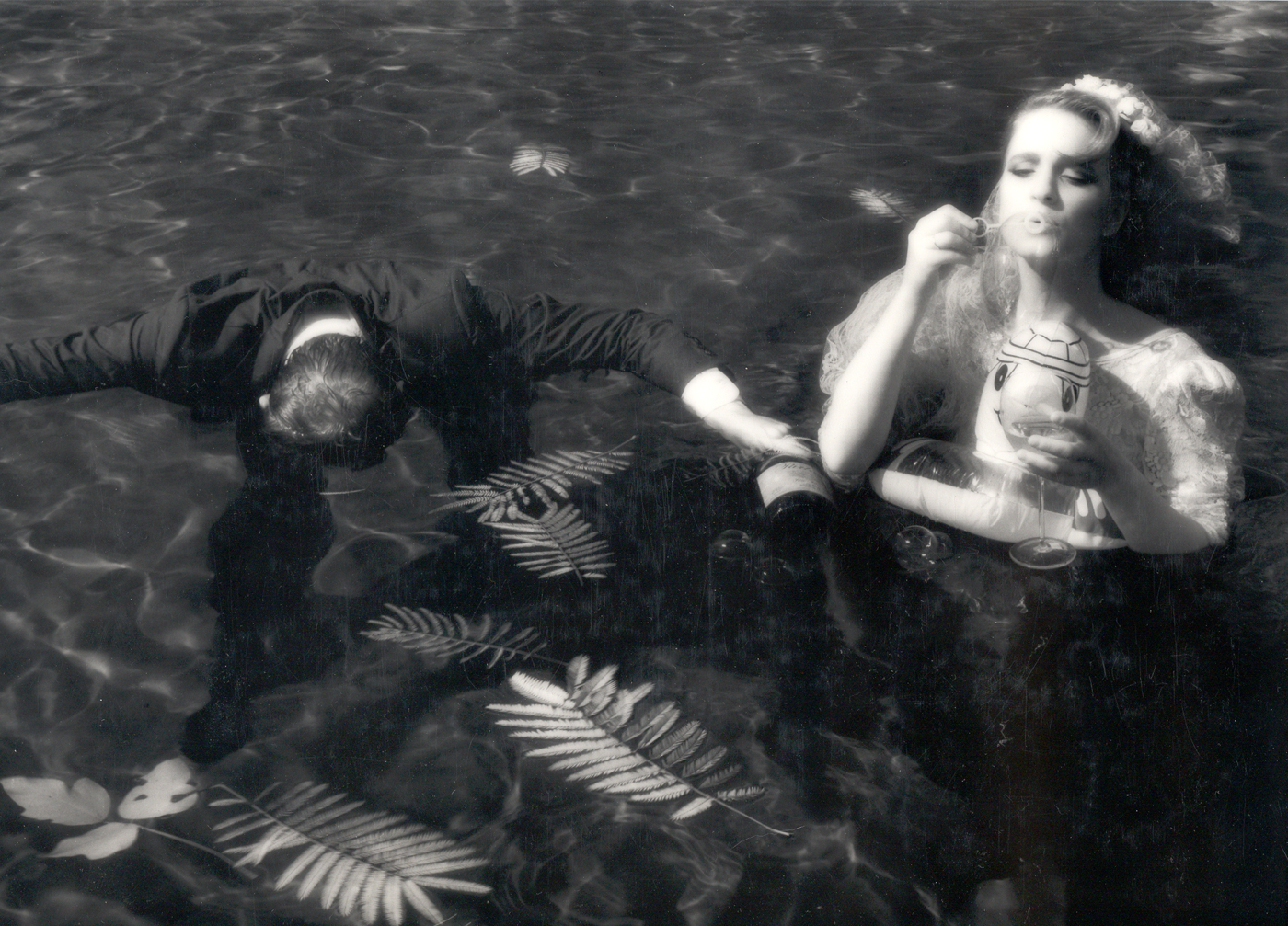 Man and woman in a pool. Man is face down in the water. Girl in white dress has flotation ring horse around her. She blows Bubbles.