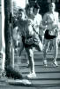Woman doing strecting excercises against telephone pole while runners pass by.
