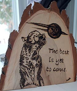 Image of a pup wolf howling at the moon with a saying: the best is yet to come, burnt on a piece of wood.