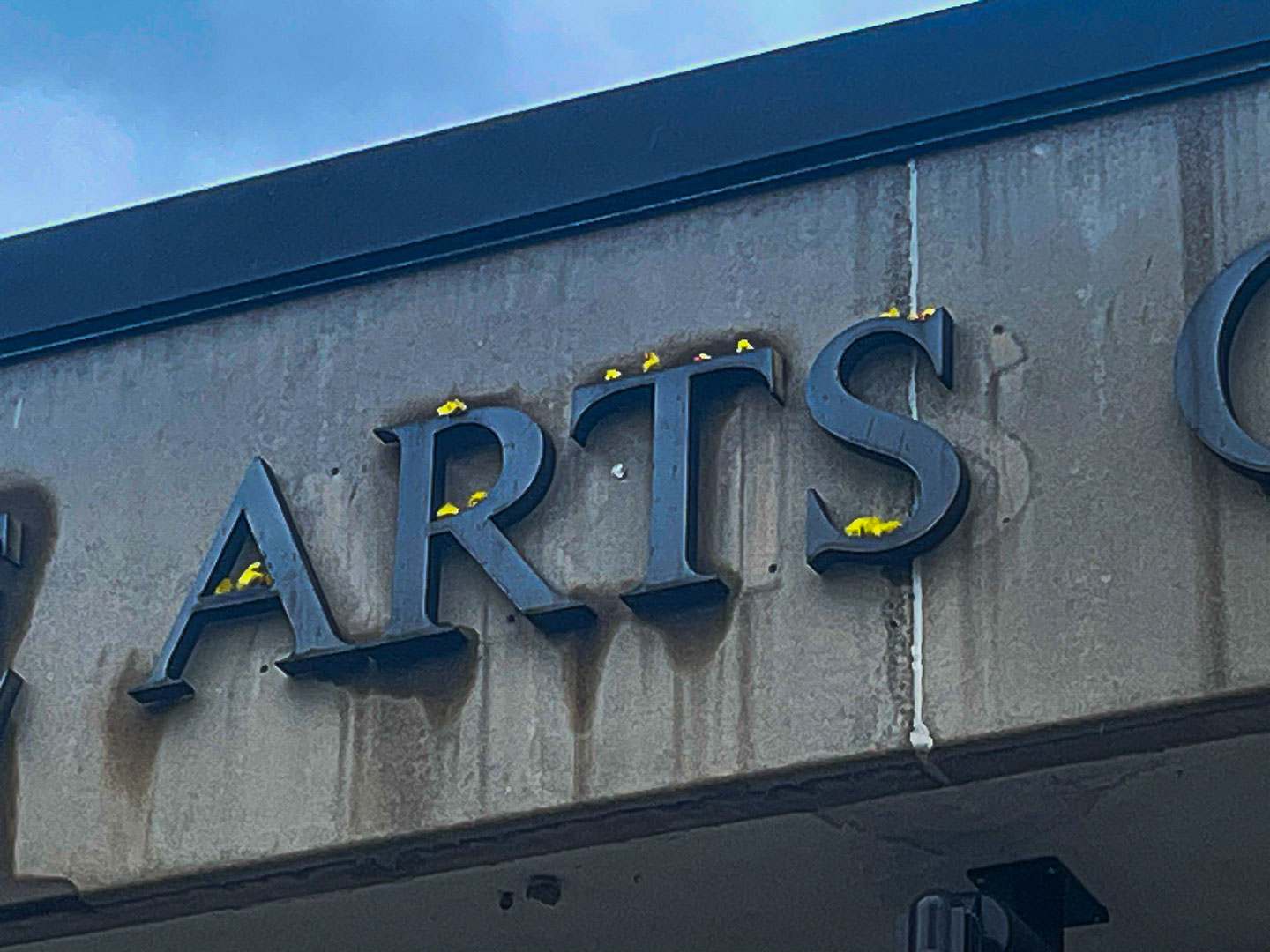 The image features a sign displaying the word ARTS, where small stuffed animal chickens in yellow are positioned on the letters. The sign is mounted on a building, with a weathered gray background that contrasts against the bright yellow of the chickens. The overall scene is set under a blue sky, enhancing the visibility of both the sign and the playful decorations.