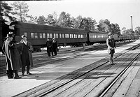 PARK RANGER LOY (TEX) WORLEY STANDING BY RR TRACKS. TRAIN AT GRCA STATION BEHIND. PARK VISITORS. CIRCA 1930 NPS PHOTO