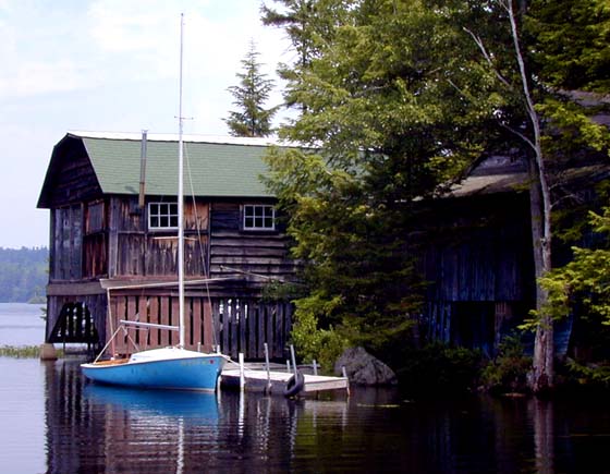 An old boothouse on a lake in New Hampshire with a blue sailboat tied up to a dock