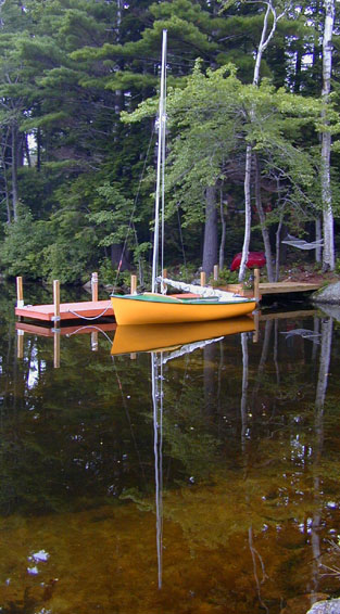 A yellow sailboat tied up at a dock on a lake with it's image reflected on the surface.